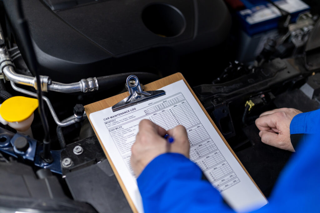 Mechanic reviewing maintenance logs while inspecting a vehicle’s engine in an auto workshop.
