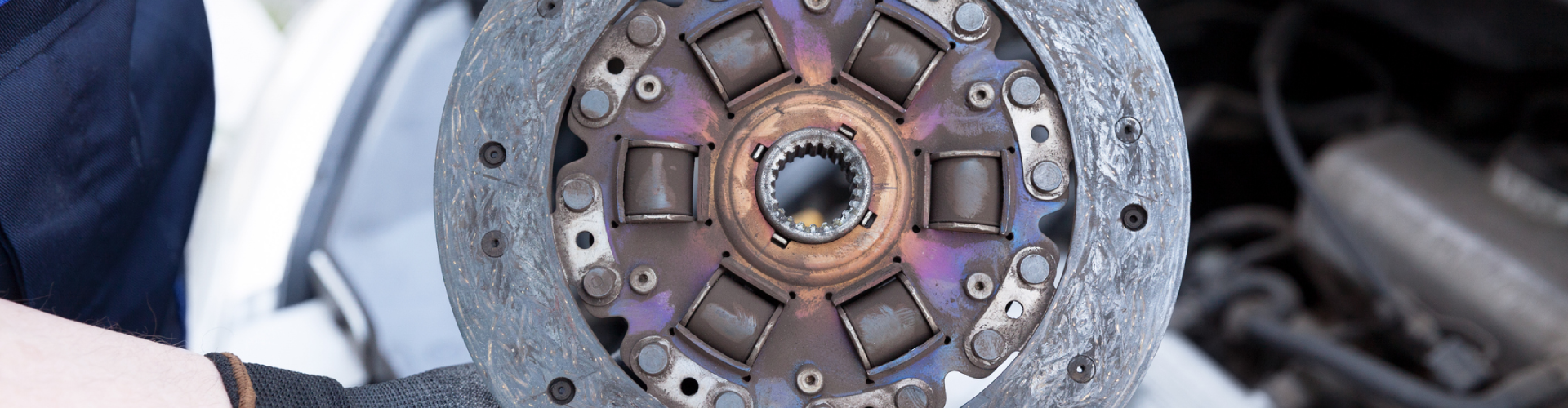 An auto mechanic wearing protective work gloves holds an old clutch disc over a BMW engine.