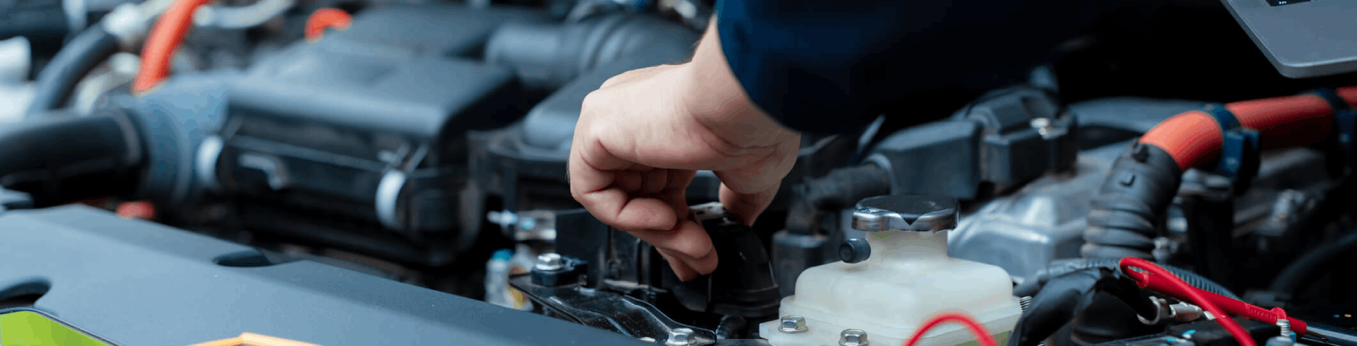 Automotive technician inspects and repairs a car’s electrical components. He holds a laptop in one hand.